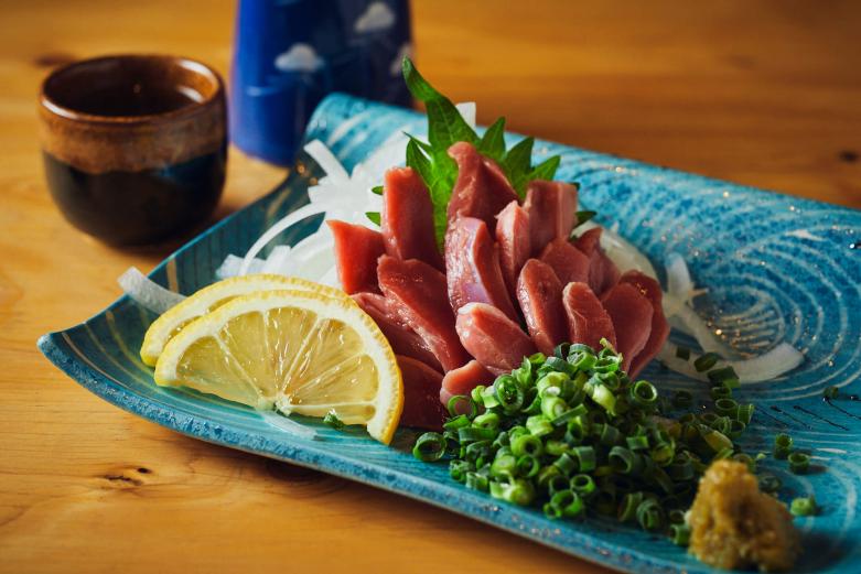 <span style="font-size:12px;"><font color="ffffff">「ズリ刺し」770円。<br> <br> 「大分では、砂肝のことを『ズリ』と呼びます」と教えてくれた伊東さん。さっそく柚子胡椒とレモン、ネギと合わせていただきます。シャキシャキした歯ごたえは新鮮な証拠です。<br> <br> 「鶏レバ刺し」「鶏ムネ刺し」（ともに770円）、「生ユッケ」（1078円）などの刺身メニューがあるなかで、「ズリ刺し」を選ぶ人って結構ツウなのでは？<br> <br> 「そうですね、独特のコリコリ食感好きの方に人気です。歯ごたえはあるけどめちゃくちゃかたくもなく、初めての人には『こんなん食べれるんや！』と驚かれます（笑）」<br> <br> まさにキモ好きにはたまらない一品。<br> <br> 肉刺しといっても完全な生食ではなく、刺身用鶏肉はすべて低温調理で殺菌し塩漬けされた“生ハム状態”で中津の仲卸から仕入れているそう。つまり衛生面も安心。<br> <br> 続いて「モモのタタキ」を。備長炭で豪快に焼き上げていきます。</font></span><br><br>