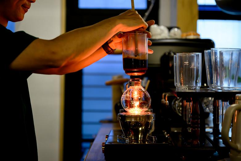 <span style="font-size:12px;">扉を開けると、ふわりと広がるコーヒーの香り。サイフォン式で丁寧に淹れられる一杯は、すっきりとした飲み口の中に、焙煎の深みをしっかりと感じられます。照明をやや落とした店内には、木の温もりが感じられるテーブルと椅子が並び、アートブックや写真集、古書などがずらり。<br>本をめくりながら、香り高いコーヒーをゆっくり味わうのも心地よいひとときです。</span><br><br>