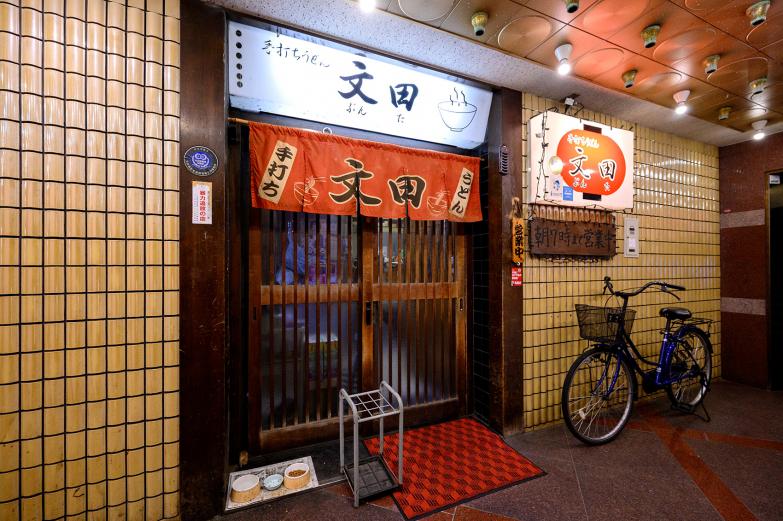 <span style="font-size:12px;"> 都町の夜を楽しんだあと、多くの人が足を運ぶのが『手打ちうどん 文田』。<br> 夜21時から朝7時まで営業しており、深夜にほっと一息つける“オアシス”のような存在です。</span><br><br>