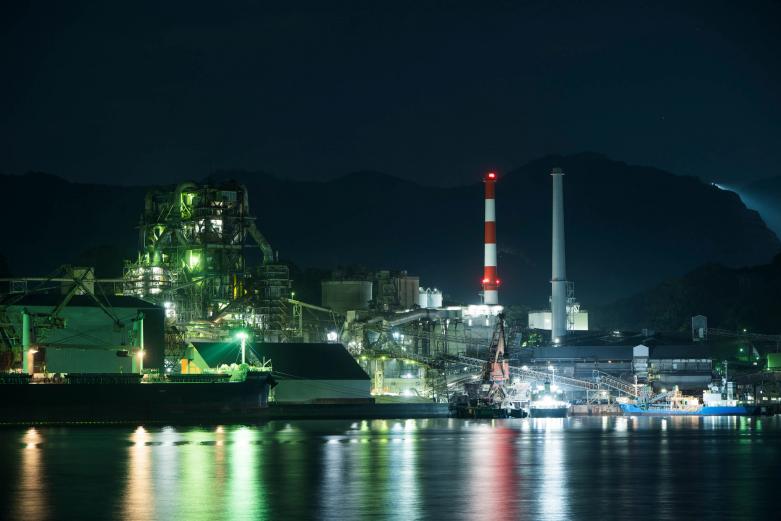 <span style="font-size:12px;"><font color="ffffff"> 工場夜景の奥に、折り重なるように鉱山の稜線がうっすらと見えます。<br> <br> 津久見ならではのセメント町の光景を満喫し、名物料理で腹ごしらえをしているうちに、次第に夜の帳が降り始めます。さあ、いよいよ工場夜景スポットへ。<br> <br> 撮影ポイントは〈つくみん公園〉と、先ほどの堅浦エリア。目の前に広がるのは、海岸沿いにずらりと立ち並ぶ工場群。点り始めた工場の灯が穏やかな水面にゆらめき、絵の具が溶け出すような幻想的な水光を描きます。</font></span><br><br>