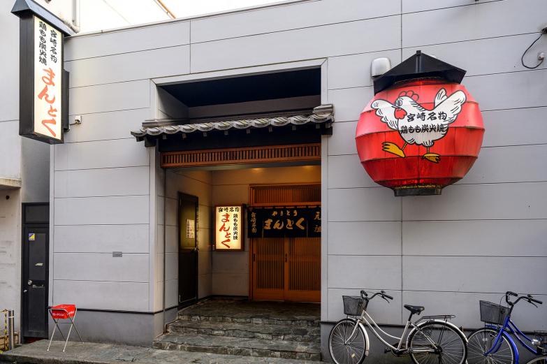 <span style="font-size:12px;"> 都町の一角にある鶏モモ焼き専門店『まんとく』。扉を開けると、炭火の香ばしい匂いとジュウッという音が店内に広がり、焼き場では炎が勢いよく上がります。<br> 都町の人気店で、オープンと同時に席は次々に埋まるので、オープン直後が狙い目です。<br> 笑い声や会話が響く店内は、まるで地元の常連と肩を並べているかのような臨場感に包まれます。</span><br><br>