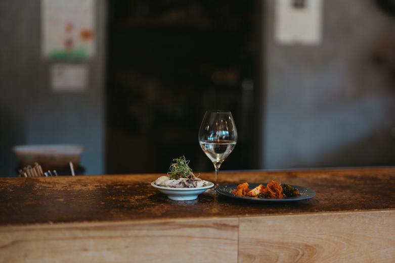 <span style="font-size:12px;"><font color="ffffff"> 自家製のキムチは常時10種前後という多彩な品揃え。どれを選べばいいの？という人にはおまかせ盛りもありますよ。この日、キムチと合わせたのが、大分にある蔵元〈浜嶋酒造〉と〈丸田酒舗〉のコラボレーションで誕生した日本酒「鷹来屋 STRIPE」です。やさしい甘さで、すいすいと飲み進められました。<br> <br> 忘れてはならないのが、店長を務める梁原仙喜(やなはらそに)さんの存在。フレンドリーな接客、そしてお酒の確かな知識と提案が心地よく、ついつい長居してしまうという人も多いようです。ここで店長を務める以前には自身で韓国料理店を営み、今でも韓国料理家としても活動。そのため、おつまみには得意のキムチもラインナップしています。「魔法使いなの!?」と思うくらいに、用いる素材は季節ごとにがらりと変わるのに、梁原さんのキムチはどれもが記憶に残る味わい。日本酒にはもちろん、ワインにもバシッとハマるキムチ。ぜひお好みの酒と一緒に楽しんでみてください。<br> <br> 「うちでしか飲めないお酒が多いのは蔵元さんとの関係があるからこそですね。毎年、蔵元さんへお酒づくりのお手伝いに行きます。そうやって交流し、つくり手の人となりを知れば知るほど、お客様へ大切に届けたいと思うんです」<br> <br> そんな梁原さんがグラスに注ぐお酒は、人の温もりが一段と感じられました。</font></span><br><br>