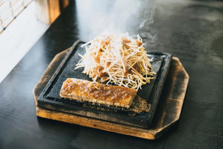 <span style="font-size:12px;"><font color="ffffff">熱々の溶岩の上でさっと炙った「まぐろステーキ」単品2050円、定食2400円。表面は香ばしく、中はしっとりレアに仕上げられ、マグロの旨みをシンプルに堪能できます。<br> <br> 鉄板ではなく溶岩プレートで焼くのがポイントだそうで、厨房では両面をさっと炙る程度にとどめます。溶岩の上でじっくり熱が伝わっていくので、食事をしながら好みの火入れ加減のマグロが楽しめるという趣向。<br> <br> 「最初はレアで、その後好みのタイミングでもやしの上に避難させちゃえば、熱々のまま堪能できますよ。味つけはシンプルなニンニク醤油と、さっぱり和風のカボスソースもご用意しています」と、簑部さん。<br> <br> つけ合わせのもやしも、マグロ同様しっとり＆カリカリと火入れによる食感の違いが楽しめて、マグロとソースの旨みをたっぷり吸ってグングン箸が進みます。</font></span><br><br>